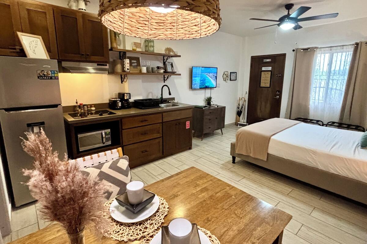 A compact hotel room with a kitchenette, a bed, a television, and a ceiling fan.
