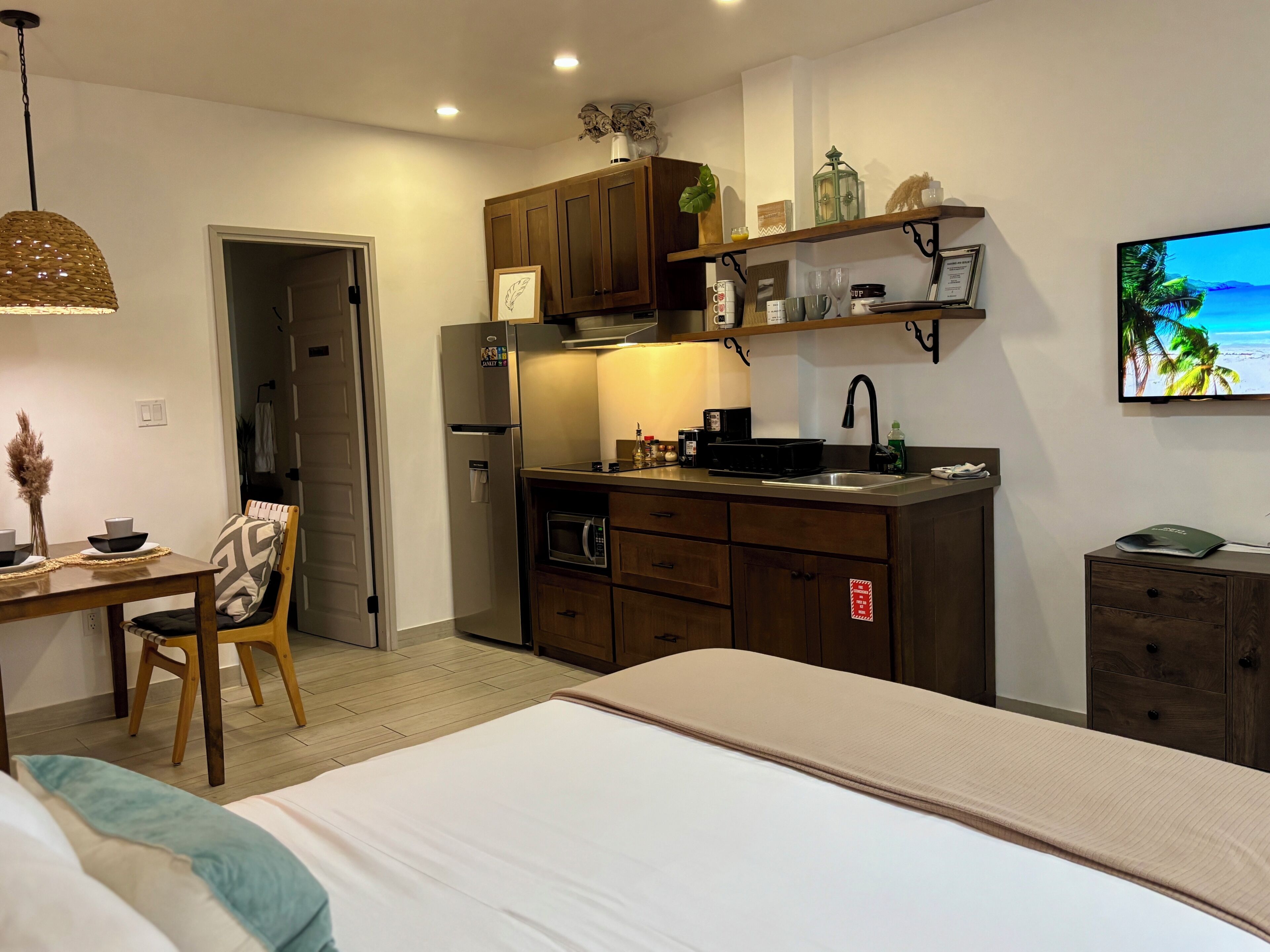 A compact hotel room with a kitchenette, a bed, a dining table with chairs, and a TV mounted on the wall.
