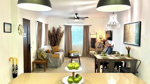 A modern living room with a dining area, featuring a sofa, a coffee table with decorative items, a ceiling fan, and a chandelier. - Stylish Apartments in Belize City (Belize City)