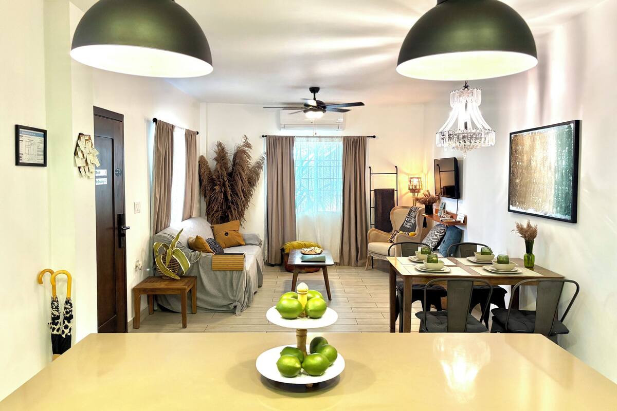 A modern living room with a dining area, featuring a sofa, a coffee table with decorative items, a ceiling fan, and a chandelier.