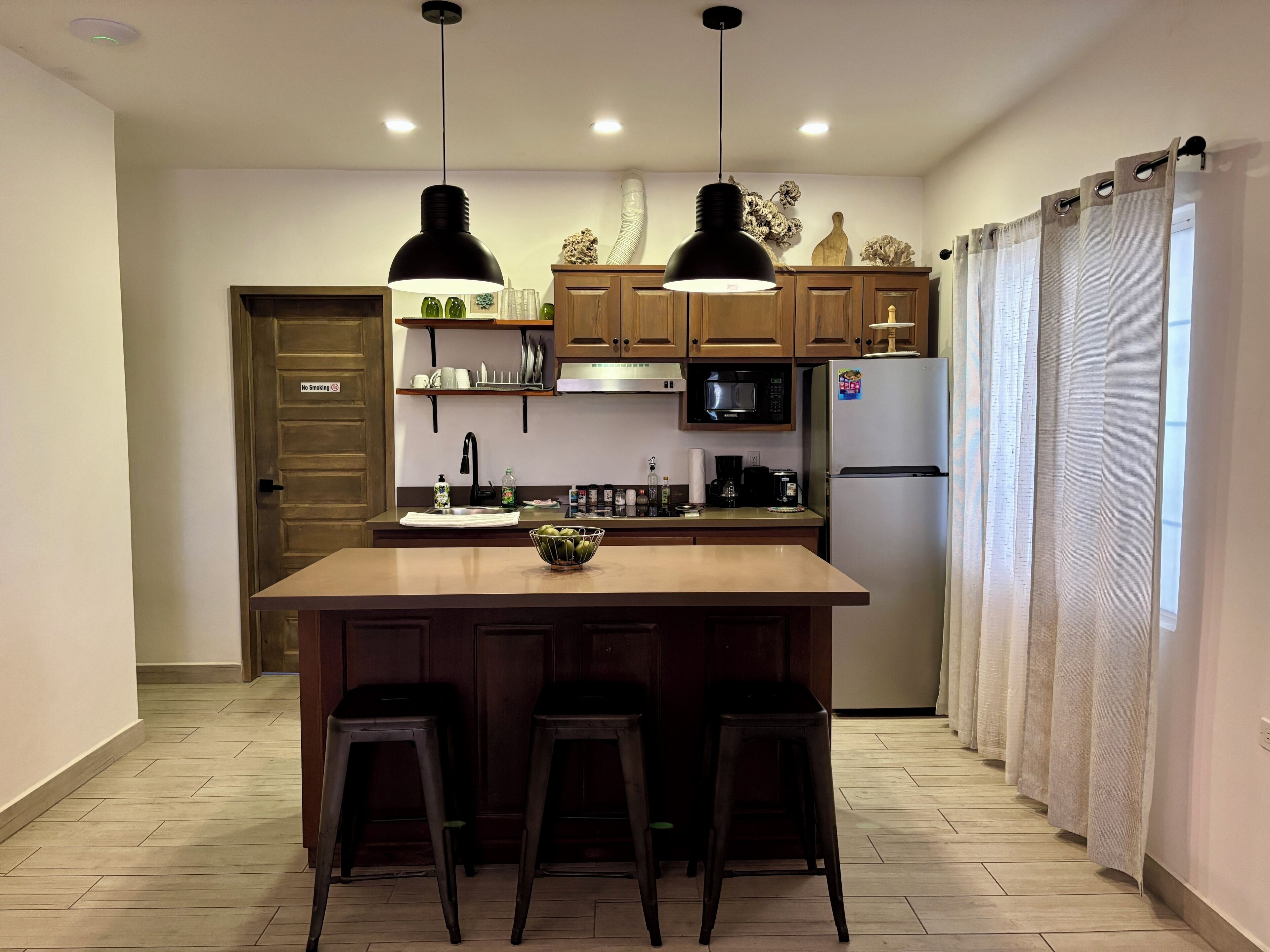 Cocina con isla central, taburetes de bar, microondas, refrigerador e iluminación colgante.