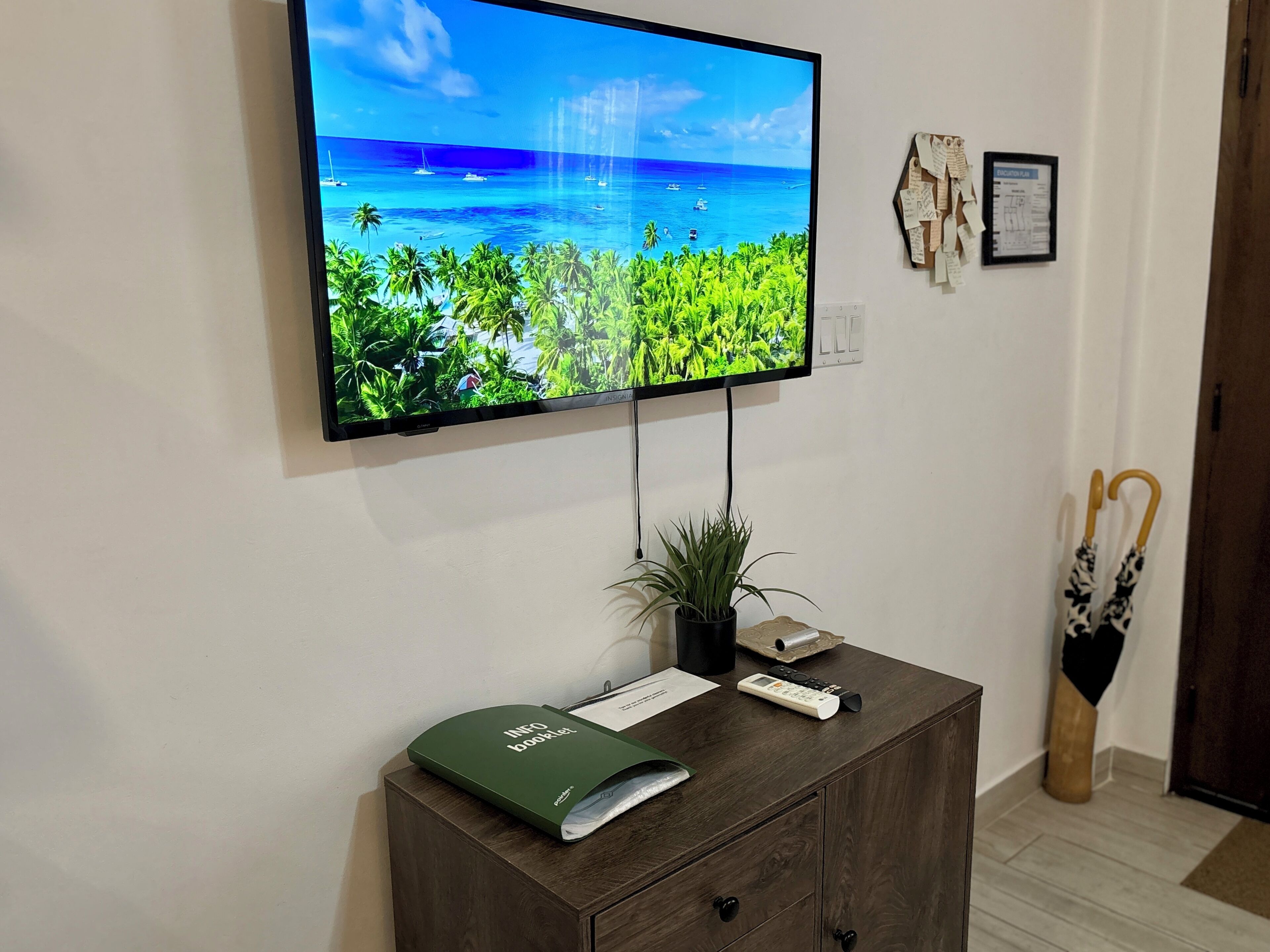 Un televisor de pantalla plana montado en la pared mostrando una playa tropical, un mueble de madera con una carpeta verde, una planta y un control remoto.