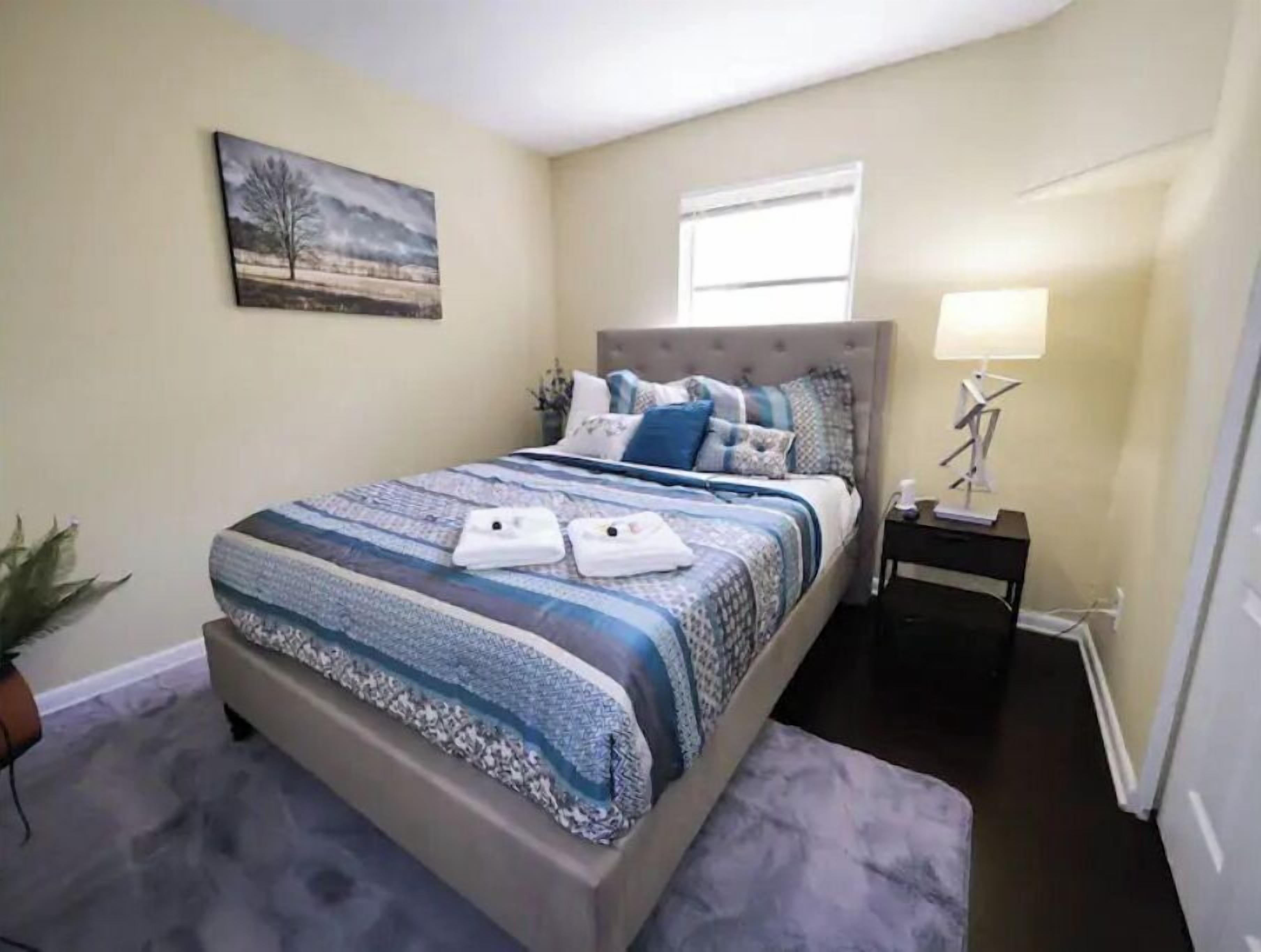 A bedroom with a bed, a nightstand, a lamp, a painting, and a potted plant.