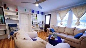 Un salon avec un canapé beige, un fauteuil bleu et une table basse. On y trouve des étagères ornées d’objets décoratifs et un ventilateur de plafond.