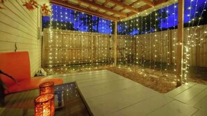 Un espace extérieur abrité, agrémenté de guirlandes lumineuses, d’une clôture en bois et d’un coin salon avec une table et des chaises.