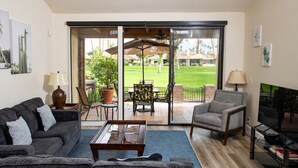 A living room with a sofa, armchair, coffee table, and a TV on a stand. There is a sliding glass door leading to a patio with outdoor furniture and a lawn. - Palm Desert Private Retreat  (Palm Desert)