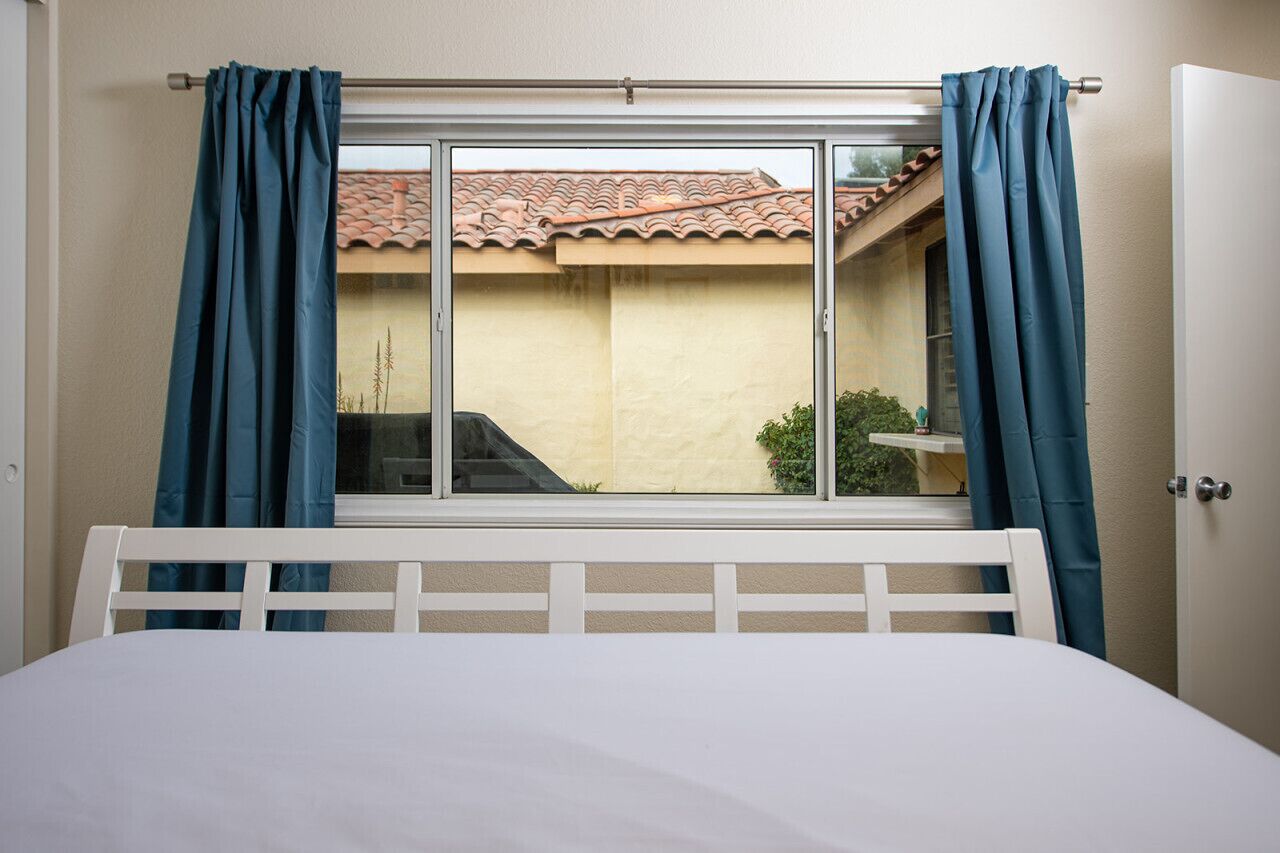 A bedroom with a bed, a window with curtains, and a view of a building with a tiled roof.