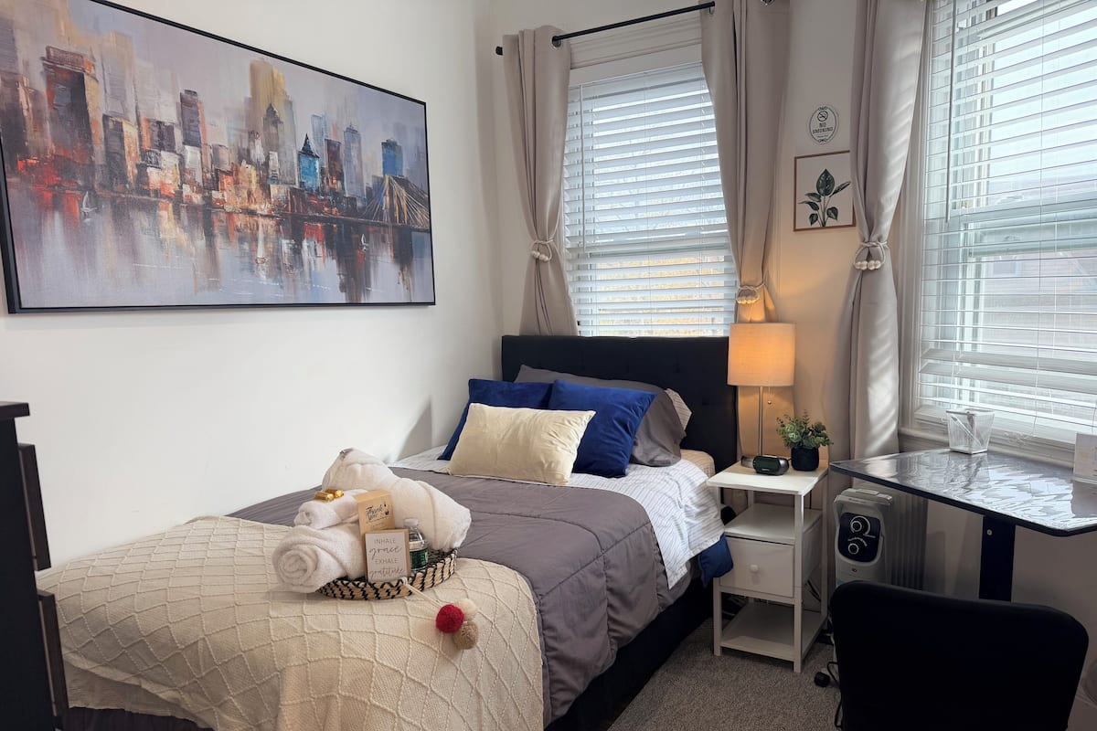 A neatly arranged bedroom with a bed, bedside table, lamp, and a cityscape painting on the wall.