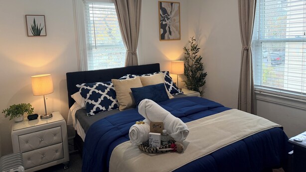 A hotel room with a bed, bedside tables, a lamp, a plant, and a window with curtains. - Cozy Private Share Room (Englewood)