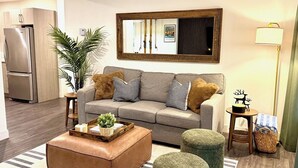 En moderne stue med en grå sofa, en brun læder skammel og to grønne puf. Et træbord med en plante står foran sofaen. Et spejl hænger på væggen over sofaen, og en lampe står på et sofabord.