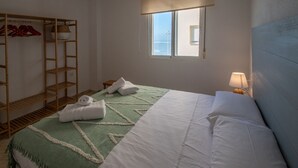 A neatly arranged bedroom with a bed, a wooden wardrobe, a window with a view of the sea, and a bedside lamp. - Apartment Tribalia - Cadiz beaches (Cádiz)