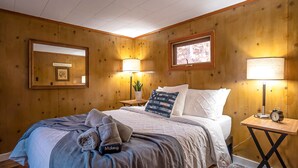 A wooden-paneled room with a bed, two bedside lamps, a framed picture, and a small table with an alarm clock.