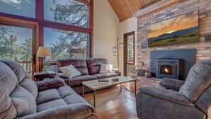 A spacious living room with a large fireplace, leather sofas, and a view of the outdoors through large windows. - Trailheads Steps Away + Stunning Views! (Pine)