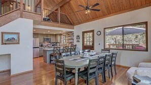 A spacious dining area with a high ceiling and wooden beams, a kitchen in the background, and a large window with a view of trees. - Trailheads Steps Away + Stunning Views! (Pine)