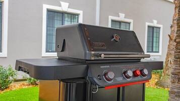 A black outdoor grill with a digital display and control knobs, positioned in front of a house with a stone pillar and a well-maintained lawn.