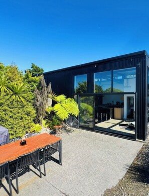 A modern house with a glass door leading to a kitchen, surrounded by a garden with various plants and a dining area with a table and chairs. - Coastal Calm | Luxe Stay with Views, Bath & Fire. (Mapua)