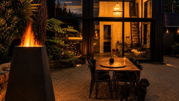 A modern two-story house with a patio area featuring a table and chairs, surrounded by plants and a fire feature. - Eco Lux Mapua | Inlet Views, Outdoor Bath & Fire (Mapua)