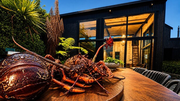 A lobster on a wooden table with a modern building in the background. - Coastal Calm | Luxe Stay with Views, Bath & Fire. (Mapua)