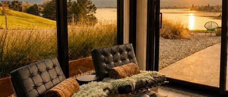 Een modern interieur met een groot raam met uitzicht op een meer en bergen, met een zithoek met een bank en voetenbankje.