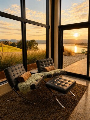 A modern interior with a large window overlooking a lake and mountains, featuring a lounge area with a sofa and ottoman. - Eco Lux Mapua | Inlet Views, Outdoor Bath & Fire (Mapua)