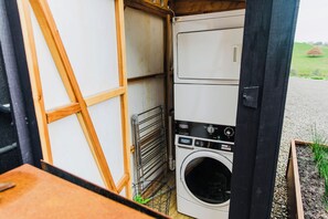 A compact laundry room with a washing machine and dryer, a metal drying rack, and a wooden door. - Coastal Calm | Luxe Stay with Views, Bath & Fire. (Mapua)
