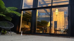 A modern building with large glass doors, a seating area with chairs, and a visible interior space with a hanging light fixture. - Coastal Calm | Luxe Stay with Views, Bath & Fire. (Mapua)