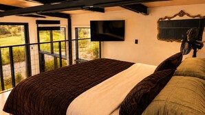 A bedroom with a large bed, a television mounted on the wall, and a balcony with a view of greenery. - Coastal Calm | Luxe Stay with Views, Bath & Fire. (Mapua)