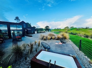 A modern outdoor patio with a hot tub, seating, and a view of the countryside. - Coastal Calm | Luxe Stay with Views, Bath & Fire. (Mapua)