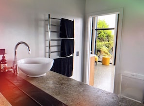 A bathroom with a white sink, a towel rack with black towels, and a view of a garden through an open door. - Coastal Calm | Luxe Stay with Views, Bath & Fire. (Mapua)