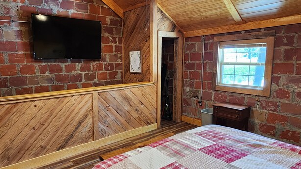 A room with a brick wall, a wooden headboard, a flat-screen TV mounted on the wall, a window with a view, and a bed with a checkered quilt.