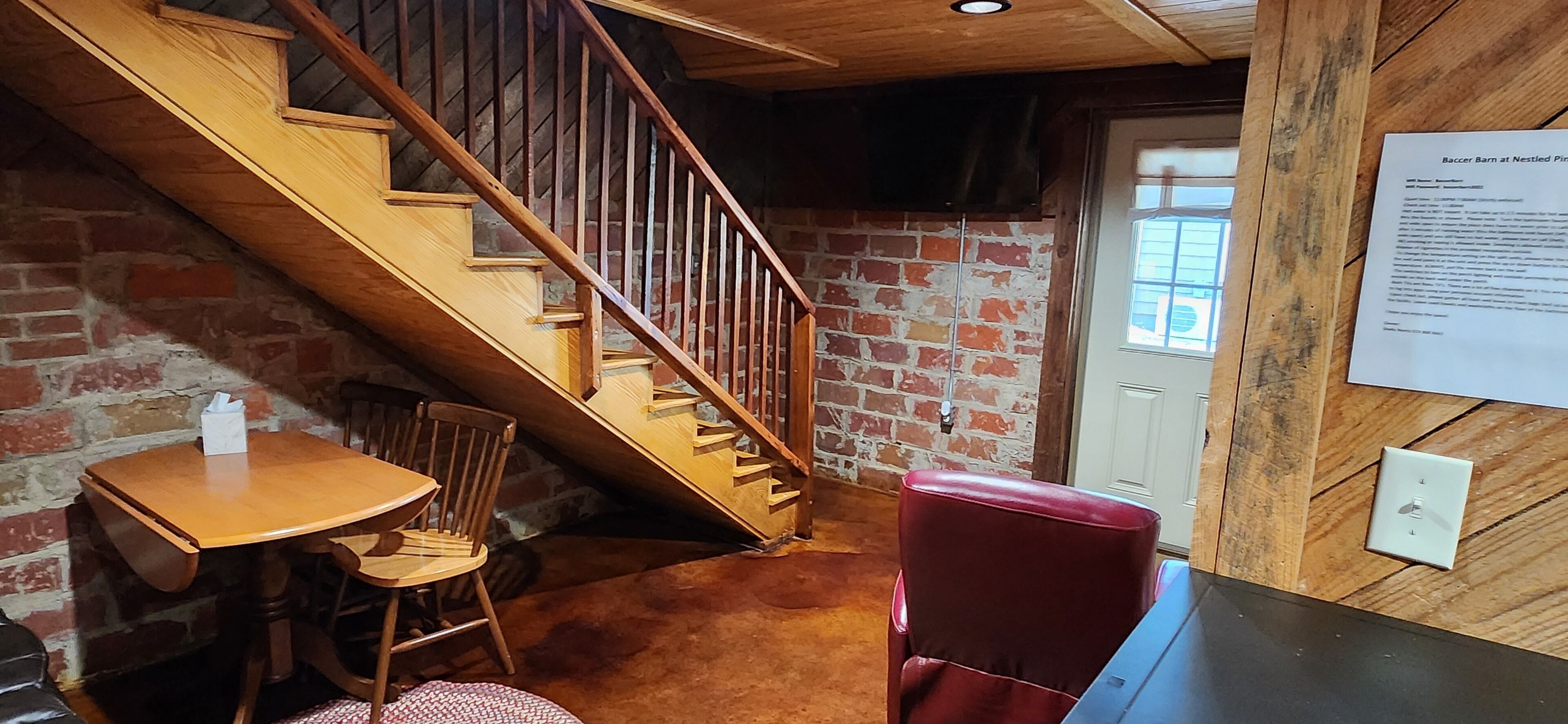Una escalera de madera que lleva a una habitación con pared de ladrillo, una mesa con sillas y una butaca roja.