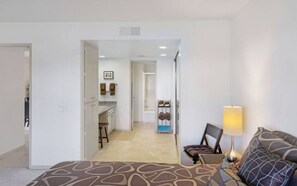 A hotel room with a bed, a desk, a chair, a lamp, and a view of the kitchen and bathroom.