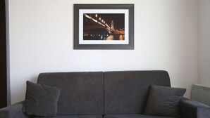Un canapé gris avec deux coussins, placé contre un mur blanc, et au-dessus, un tableau encadré représentant un pont de nuit.