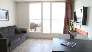 Une pièce moderne avec un canapé gris, une petite table sur laquelle repose un magazine, un téléviseur fixé au mur et un balcon offrant une vue agréable.