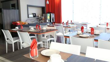 A modern dining area with white chairs, tables, and red napkins. There is a coffee machine and a counter with a refrigerator in the background.