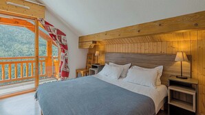 A bedroom with a wooden headboard, a bed with white bedding, a nightstand with a lamp, and a view of a forest through a balcony.