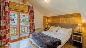 A bedroom with a wooden headboard, a bed with white linens, a bedside table with a lamp, and a view of a stone building through a glass door.