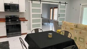 A compact kitchen with a microwave, oven, and white cabinets. A dining table with chairs is in the foreground, and a sofa is in the background. - Stylish, comfortable, and private Guest House. (Eastman)
