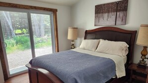 Quarto com cama de madeira, uma janela grande com persianas, um quadro na parede e uma mesa de cabeceira com abajur.