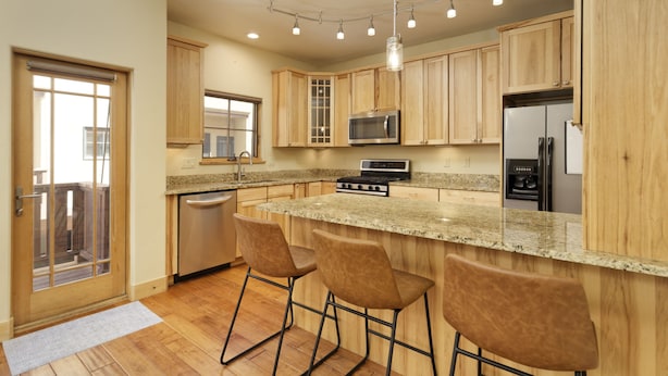 A modern kitchen with wooden cabinets, a granite countertop, a stainless steel refrigerator, and a built-in oven. There are three bar stools and a window with a view of the outside. - Excellent Location- Downtown Carbondale on Main St.! (Carbondale)
