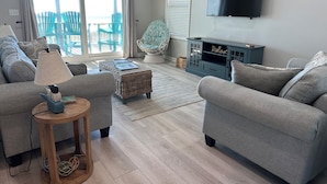 Een woonkamer met twee grijze banken, een houten bijzettafeltje, een lamp, een rieten poef, een blauwe fauteuil, een televisie en een kast met een 'Relax'-bordje.