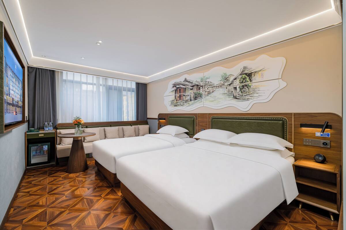 Ein Hotelzimmer mit zwei Betten, einem kleinen Tisch mit einer Vase, einer Couch, einem Fernseher und einem an der Wand befestigten Kunstwerk.