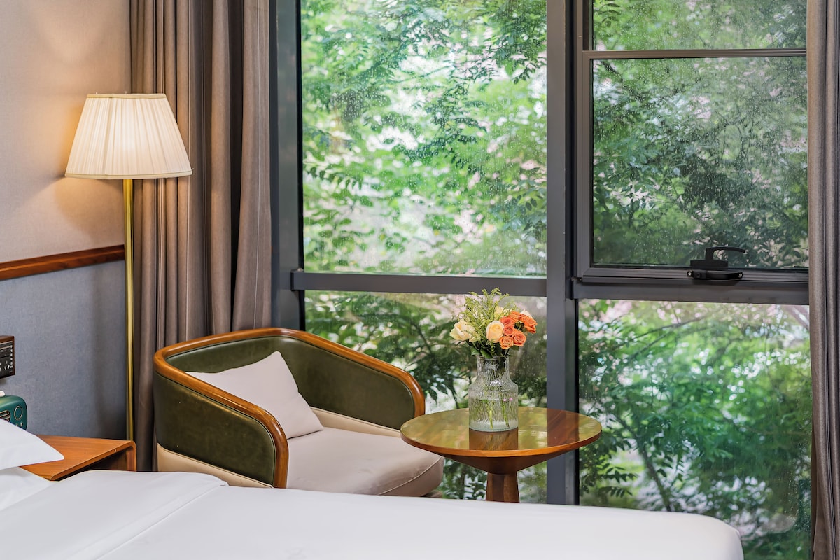 A hotel room with a bed, a chair, a table with a vase of flowers, a lamp, and a window with a view of trees.