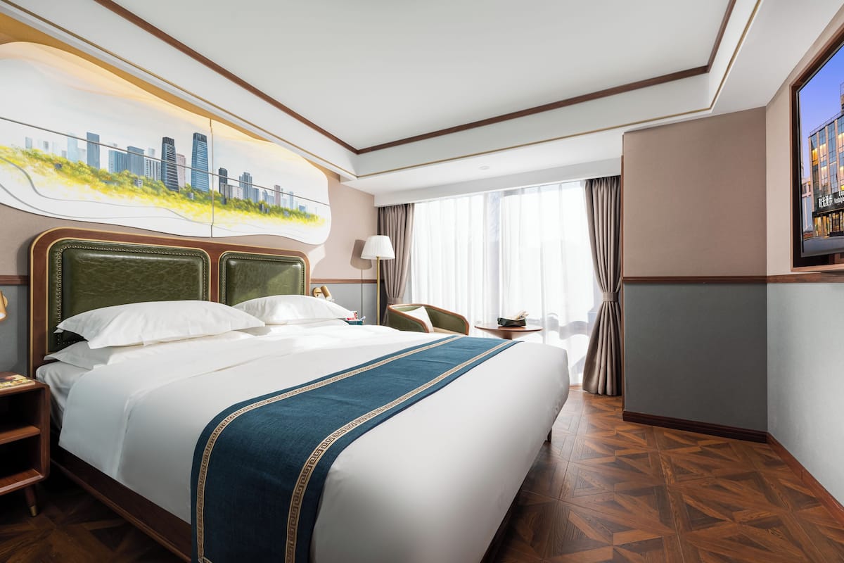 Ein Hotelzimmer mit einem großen Bett, einem Stadtbild als Gemälde, einem Schreibtisch mit Lampe und Blick auf die Stadt durch das Fenster.