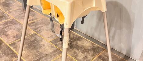 Una silla alta amarilla con base color beige, ubicada sobre un piso de baldosas con líneas amarillas.