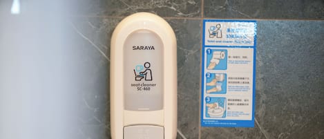 A hand sanitizer dispenser labeled 'SARAYA SC-460' and a multilingual instructional sign on a tiled wall.