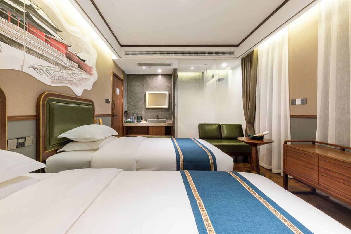 Ein Hotelzimmer mit zwei Betten, einem grünen Sofa, einem Holzkasten und einem Badezimmer mit Spiegel, sowie einer Wand mit einem großen Poster.