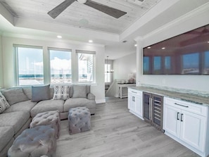 A modern living room with a sectional sofa, ottomans, a kitchen with white cabinets, and large windows overlooking a beach. - Beach Country 7 BR/ 7.5 House w/ Heated Pool (Navarre Beach)