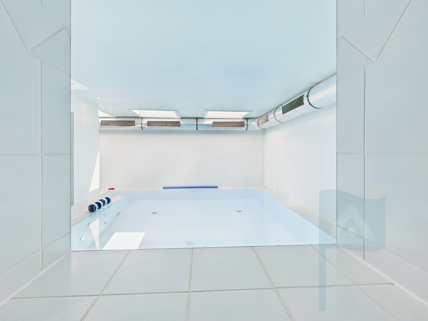 A modern, empty room with a large, rectangular pool, white tiled walls, and a ceiling with recessed lighting.