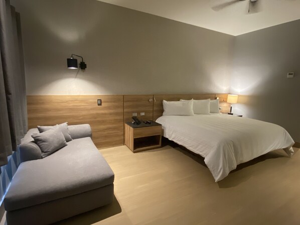 A modern hotel room with a large bed, a sofa, a nightstand, and a wall-mounted lamp.